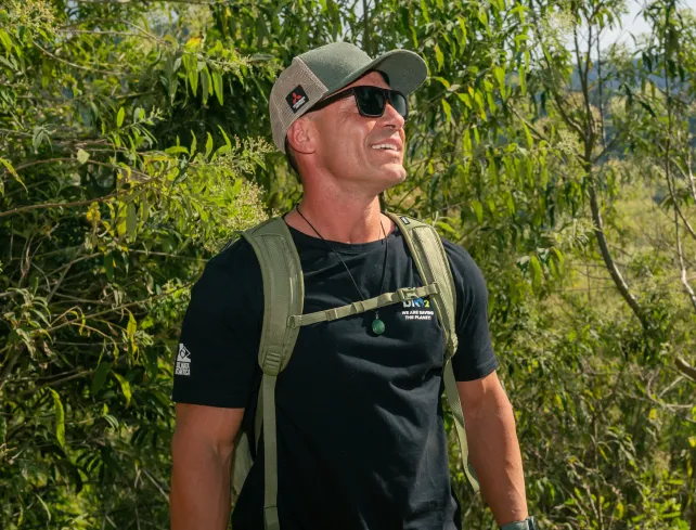 Pedro Oliva fazendo trilha Floresta biO2 olhando pra cima dando risada vestindo uma camiseta preta e mochila verde da Thule