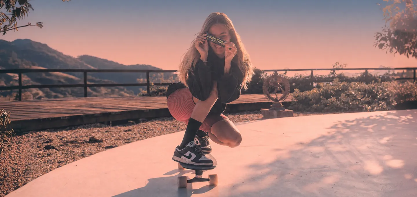 Mulher agachada sobre um skate ao pôr do sol segurando uma barra biO2 Protein na frente dos olhos