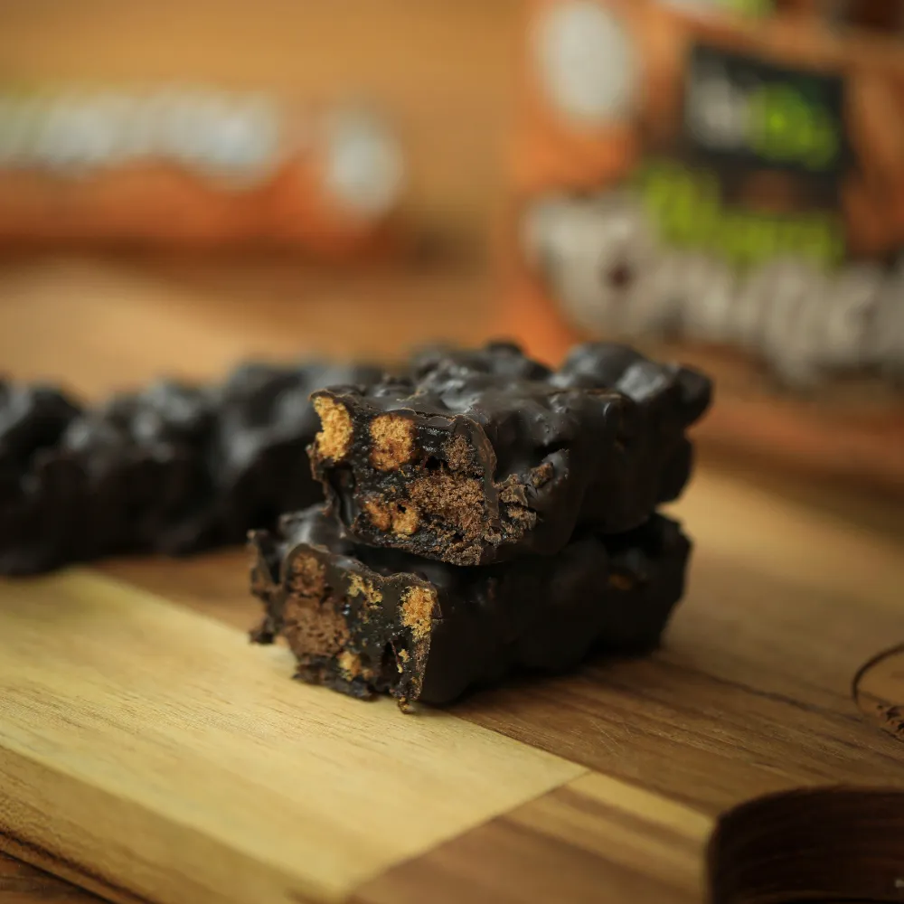 Barra de cereais crocantes biO2 cortada ao meio, vegana e sem glúten, sabor cenoura e cacau com cobertura de chocolate meio amargo, em cima de uma tábua de madeira