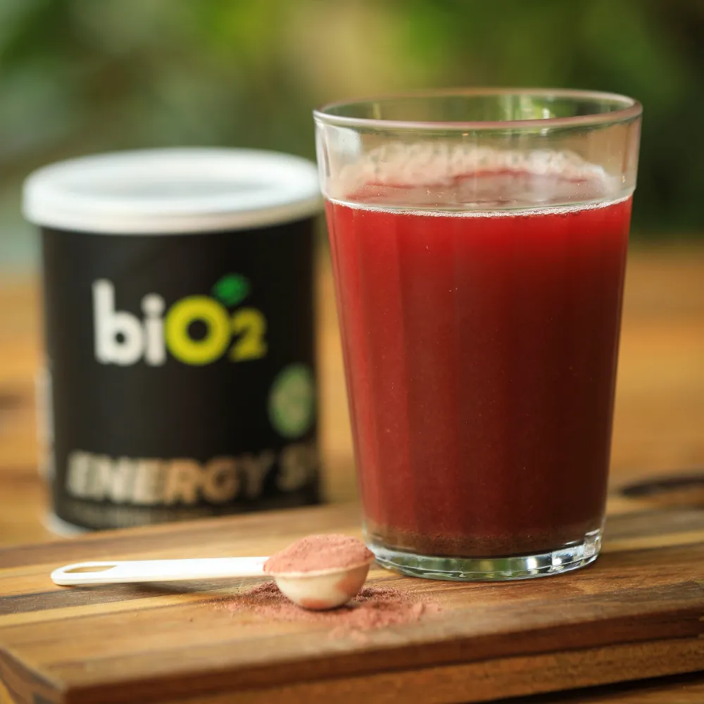 Copo de vidro com líquido vermelho, mostrando a cor do Pré-Treino em pó biO2, vegano e sem glúten, uma colher de dosagem na frente do copo com o pré-treino.