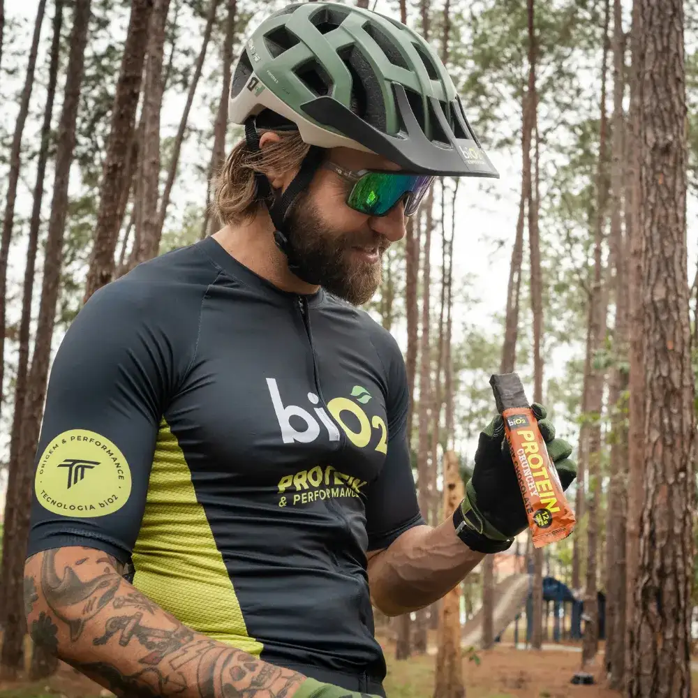 Ciclista de Mountain Bike, usando capacete e uniforme biO2 Protein & Performance, segura a Barra de Proteína Crocante biO2, vegana e sem glúten, sabor Dark Chocolate na floresta.