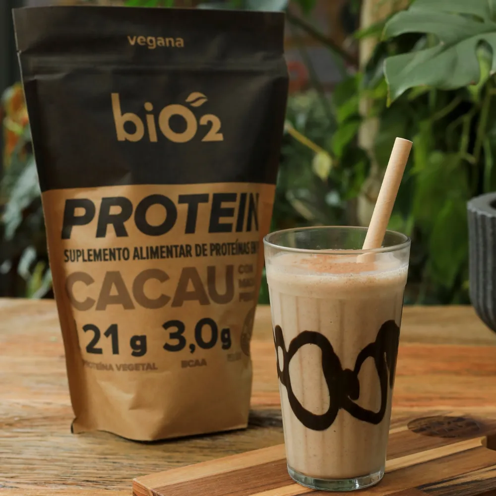 Embalagem de 908 gramas da proteína em Pó biO2, vegana e sem glúten, sabor cacau e maca peruana, ao lado de um shake proteico decorado de mesmo sabor.