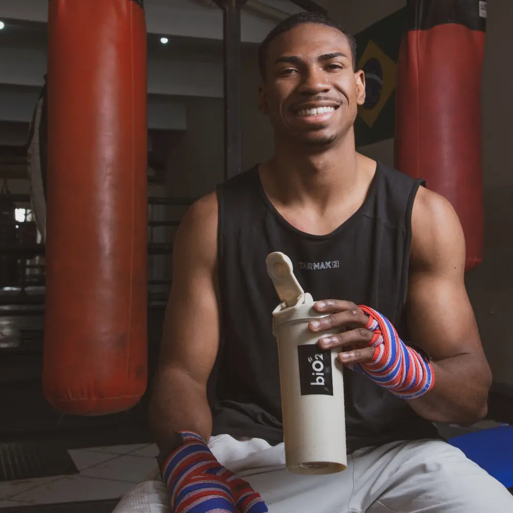 Atleta de boxe sorrindo, segurando a coqueteleira biO2 sustentável, após o treino, com saco de boxe ao fundo.
