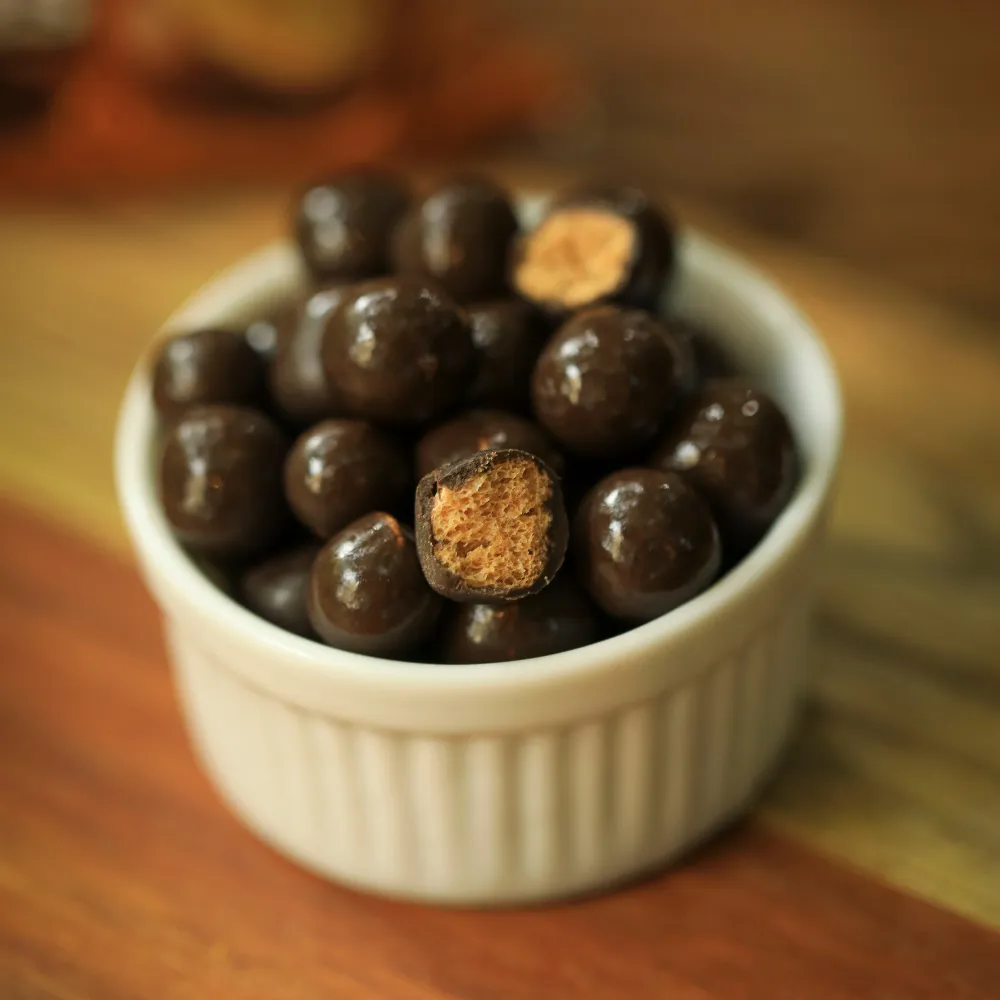 Bolinhas de cereais crocantes biO2, vegana e sem glúten, sabor cenoura com cobertura de chocolate meio amargo, em um pote de cerêmica branco com duas bolinhas cortadas ao meio.