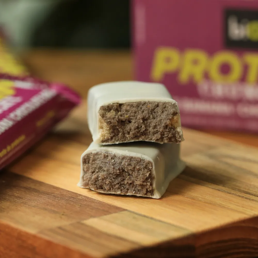 Barra de Proteína Crocante biO2, cortada ao meio, vegana, sem glúten, sabor açaí e banana com cobertura de chocolate branco, 50 gramas.