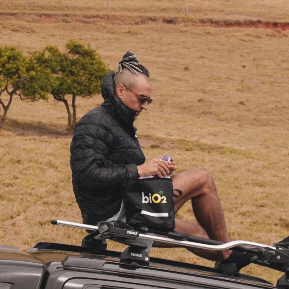 Homem sentado no rack Thule de um carro em um campo, segurando uma lancheira térmica biO2 e tirando um snack para comer durante a expedição.