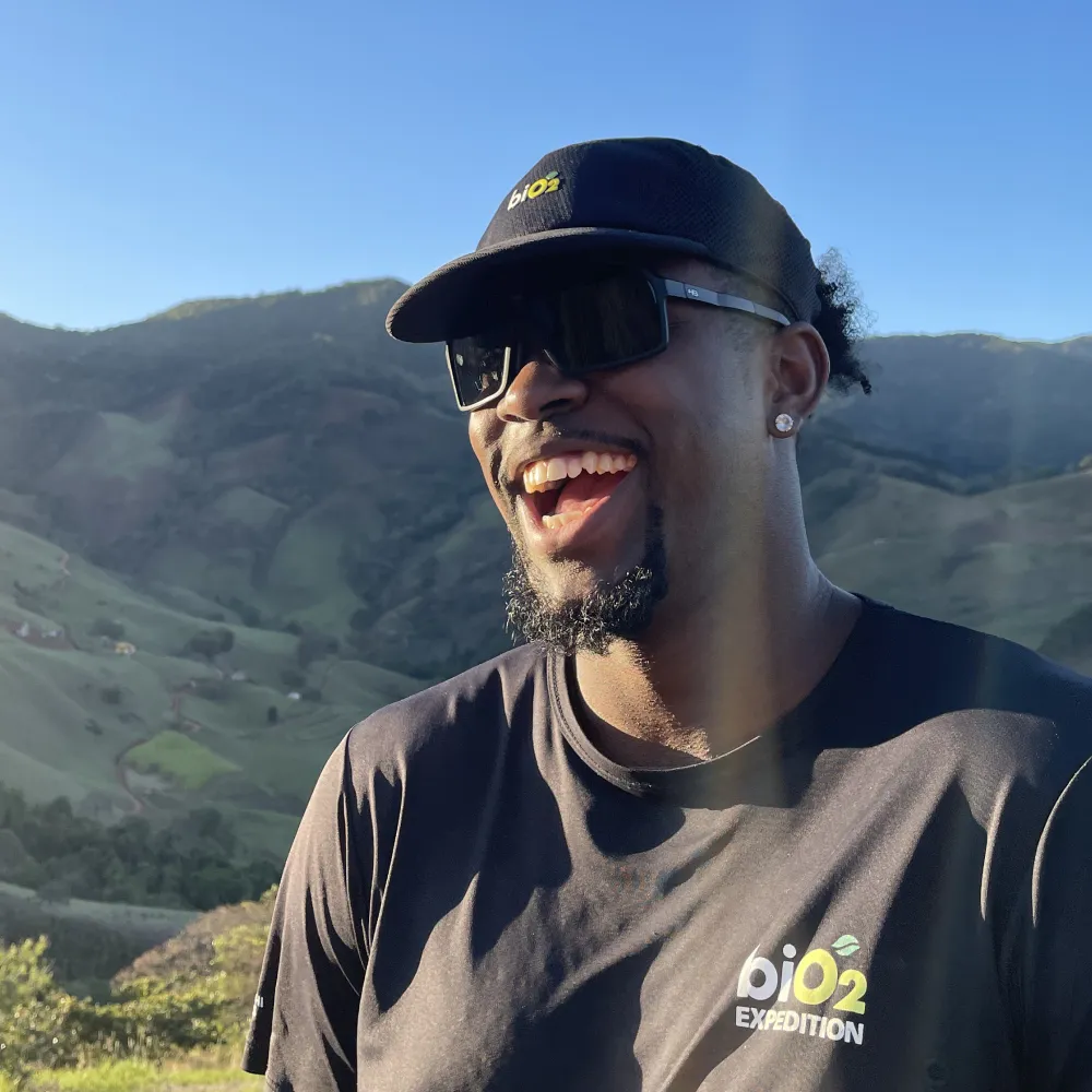 Homem com montanhas verdes ao fundo dando risada vestindo boné preto biO2 five panel aba reta, óculos e camiseta preta biO2 expedition