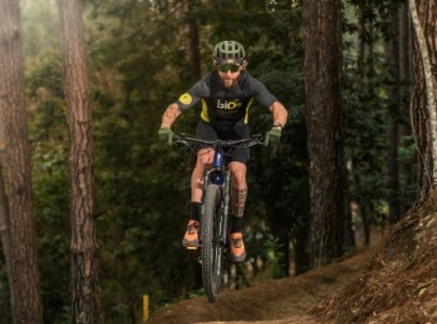 Ciclista de mountain bike saltando em uma trilha de terra em meio à floresta, vestindo uniforme esportivo da marca bio2. Ele utiliza equipamentos de segurança como capacete, luvas e óculos escuros.